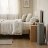 asset_12e432db1b970dfc Grey tower air purifier running beside a neatly made bed in a bright morning bedroom
