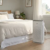 asset_4d012d59f97fec42 Winix C610 air purifier running beside a neatly made bed in a sunlit bedroom