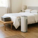 A Winix air purifier standing beside a neatly made-up guest bed with a small suitcase on a bench