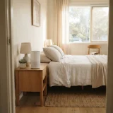 asset_987e1b2476aa5ed4 A sunlit bedroom with a plain compact air purifier standing beside the bed