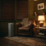 asset_9a8feda3e2f434e3 Grey tower air purifier running beside a leather armchair in a warmly lit den with closed blinds