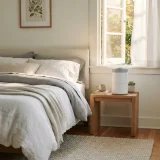 asset_a175451f59d97f8e Air purifier running beside a bed in a sunlit bedroom during pollen season