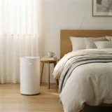 A white air purifier running beside a bed with soft morning light in a tidy bedroom.