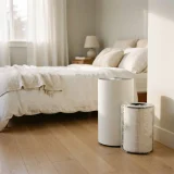 Air purifier with a filter beside a neatly made bed in a sunlit bedroom