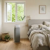 A Winix air purifier beside an unmade bed with an open window and leafy branches outside