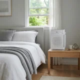 A Winix C610 air purifier running beside a bed in a sunlit bedroom with the window slightly open