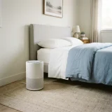 asset_dd76d191371fdfd4 Compact air purifier running on a bedroom floor beside a made bed