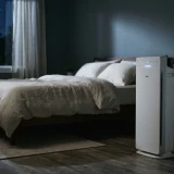 Winix AM90 air purifier glowing softly beside a made bed in a dark bedroom at night