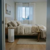Winix C545 air purifier running beside a bed in a softly lit bedroom at dawn