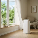 Winix AM90 air purifier beside an open window with trees visible outside on a spring day