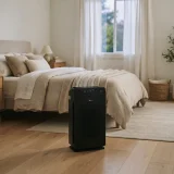 Winix 5510 air purifier running at the foot of a neatly made bed in a calm modern bedroom